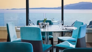 Un coin repas avec une table dressée pour quatre personnes, de grandes fenêtres offrant une vue sur la mer et les montagnes lointaines.