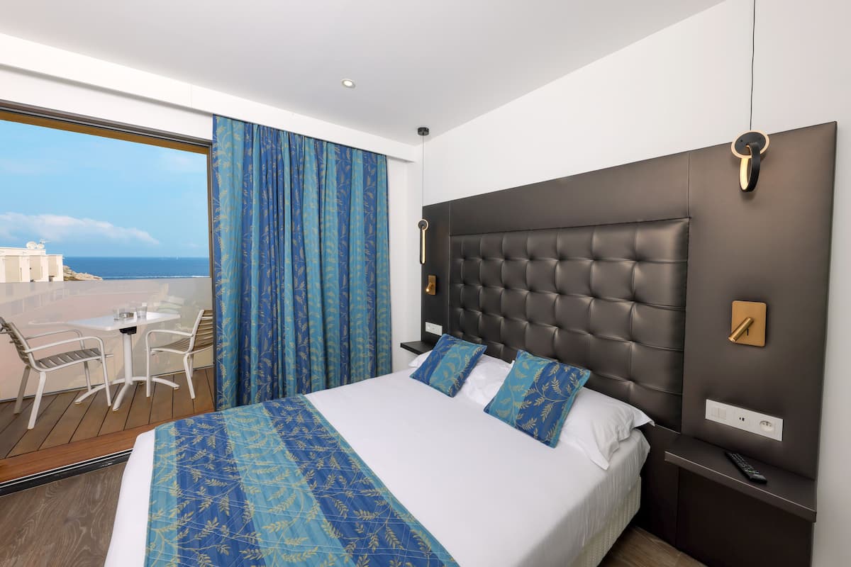 Une chambre d’hôtel moderne dotée d’un grand lit, offrant une vue sur la mer, d’un balcon avec une table et des chaises, ainsi que d’un panneau de commande mural.