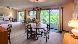 A spacious living room with a large window, a dining area with a round table, and a ceiling fan.