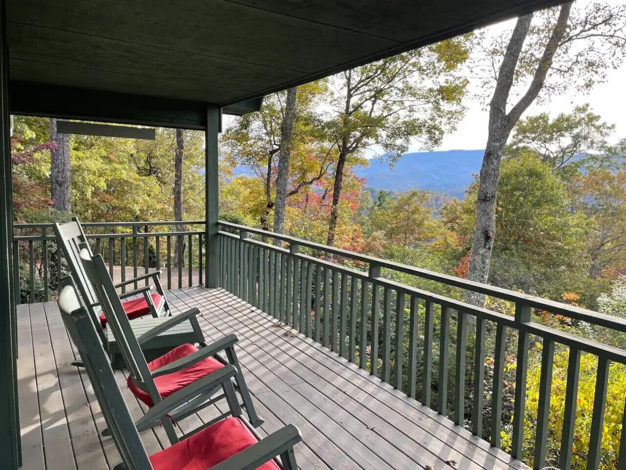 A wooden deck with a railing, two lounge chairs with red cushions, and a view of trees and mountains.