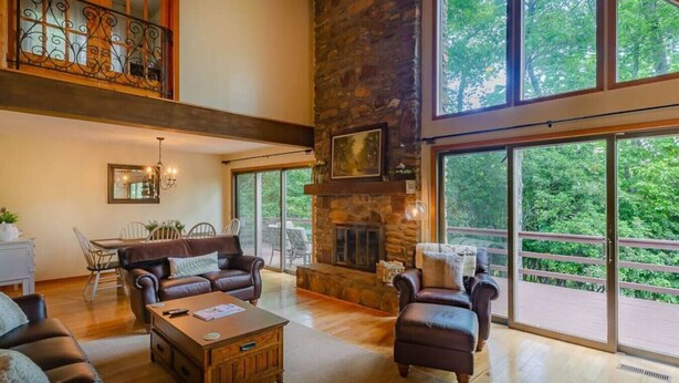 A spacious living room with a stone fireplace, large windows, and a balcony with a view of trees. - Rosewood Cottage Tucked Away in the Mountains (Franklin)