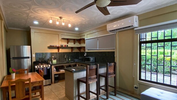 A compact kitchen with a central island, a dining table with chairs, a ceiling fan, and a large window with a view of greenery. - 2 Room Family Home in Bahia, Gated, walking distance to National Park (Bahia Ballena)