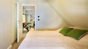 Ein Schlafzimmer mit schrägen Wänden, einem Bett mit grünen Kissen, einem Schreibtisch mit Stuhl und einer Pendelleuchte.