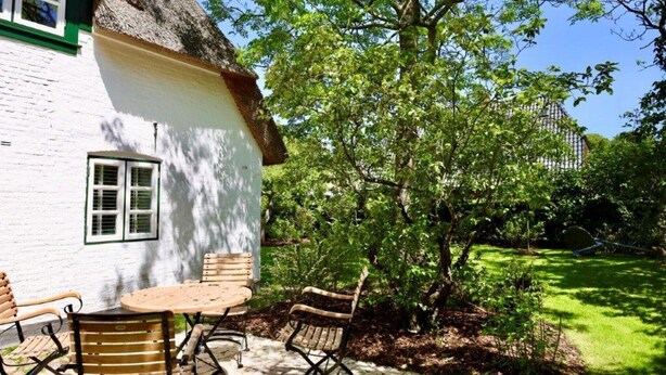 A white house with a thatched roof, a wooden table, and two chairs in a garden.
