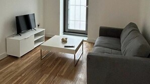 Una sala de estar moderna con un televisor de pantalla plana sobre un soporte blanco, un sofá gris y una mesa de centro blanca con un control remoto.