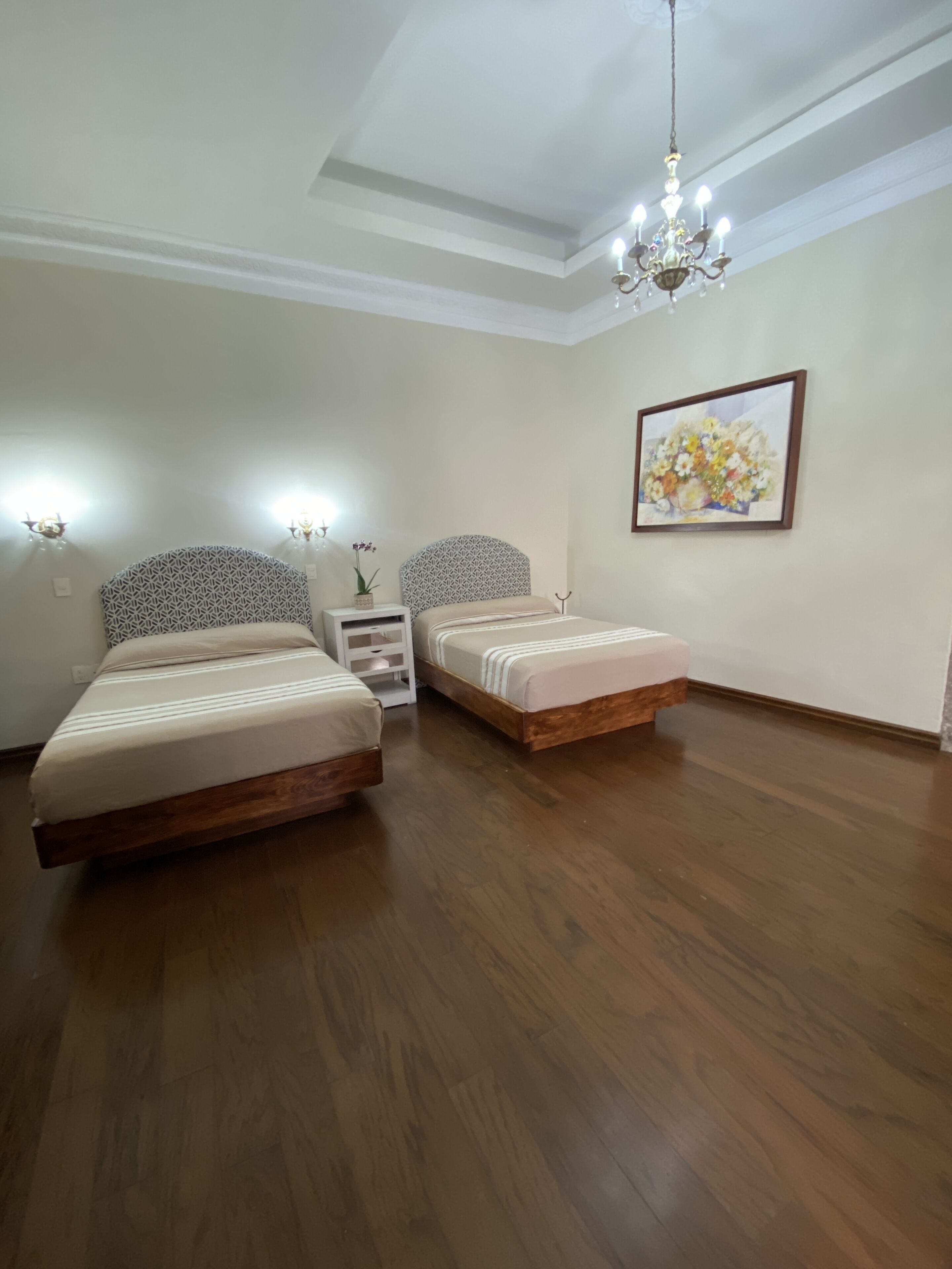 A bedroom with two beds, a chandelier, a wall-mounted picture, and a nightstand with a vase.