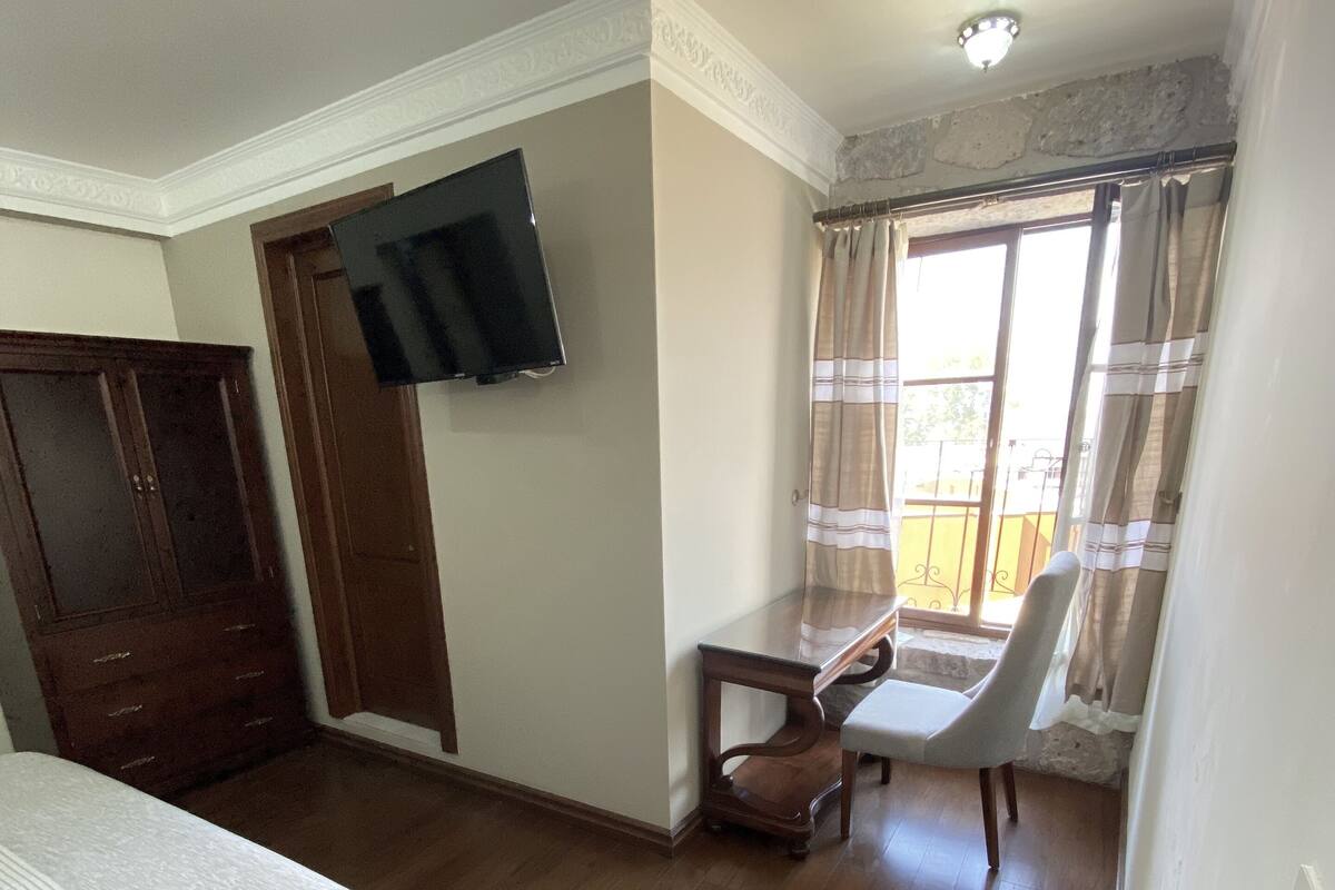 Un dormitorio con cama, televisor montado en la pared, escritorio de madera, silla y ventana con cortinas.