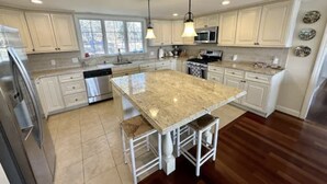 A modern kitchen with white cabinets, a large island with bar stools, stainless steel appliances, and pendant lighting. - WATER FRONT- saltwater private pond beach and water sports IDEAL for big groups! (Dennis)