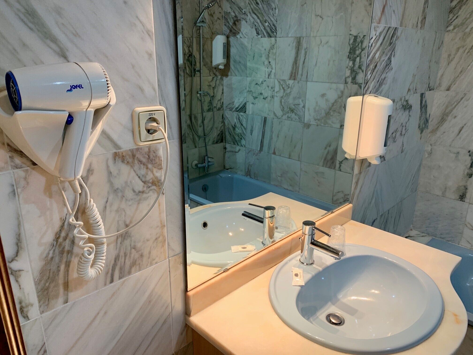 A bathroom with a marble wall, a sink, a mirror, a handheld hair dryer, and a handheld showerhead.
