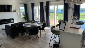 A modern kitchen with a central island, dining area with round table and chairs, and a view of a deck and lake through large windows.