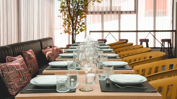Un restaurante con una larga mesa de comedor dispuesta con platos, vasos y velas. Hay sillas amarillas y un sofá negro con cojines estampados.