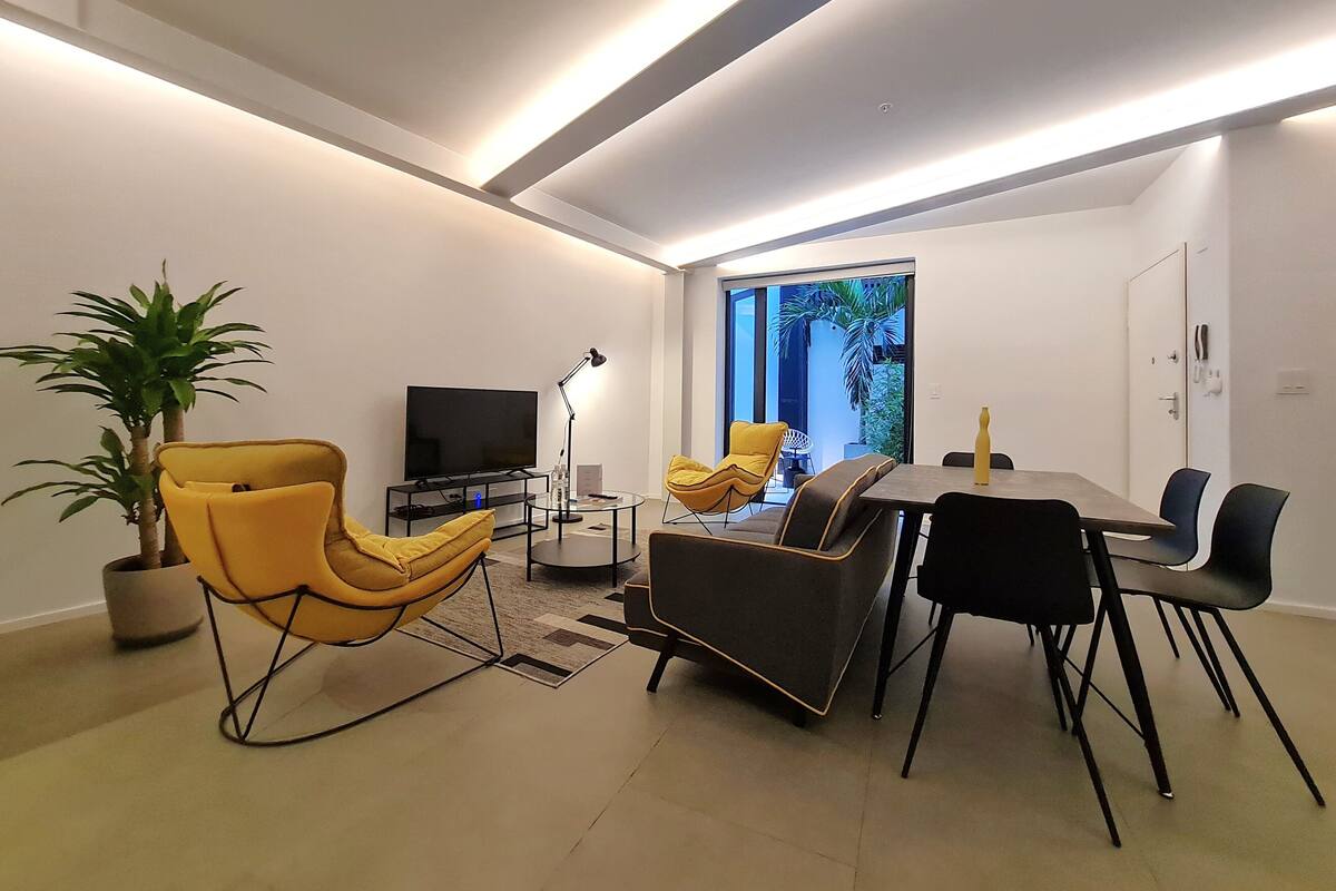 Ein modernes Wohnzimmer mit einem Flachbildfernseher, gelb-schwarzen Sesseln, einem Esstisch und einem großen Fenster mit Blick auf Grünflächen.
