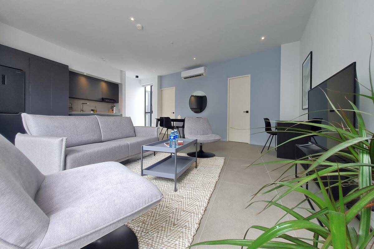 Ein modernes Wohnzimmer mit einem grauen Sofa, einem Couchtisch und einem Fernseher. Es gibt eine Küchenzeile mit dunklen Schränken und einem Flachbildfernseher.
