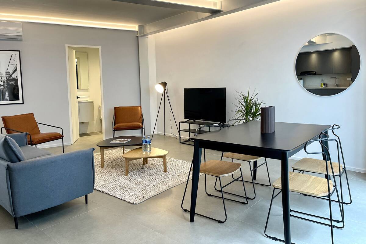 Ein modernes Wohnzimmer mit Sofa, Sessel, Couchtisch, Fernseher und einer Essecke mit Hockern.
