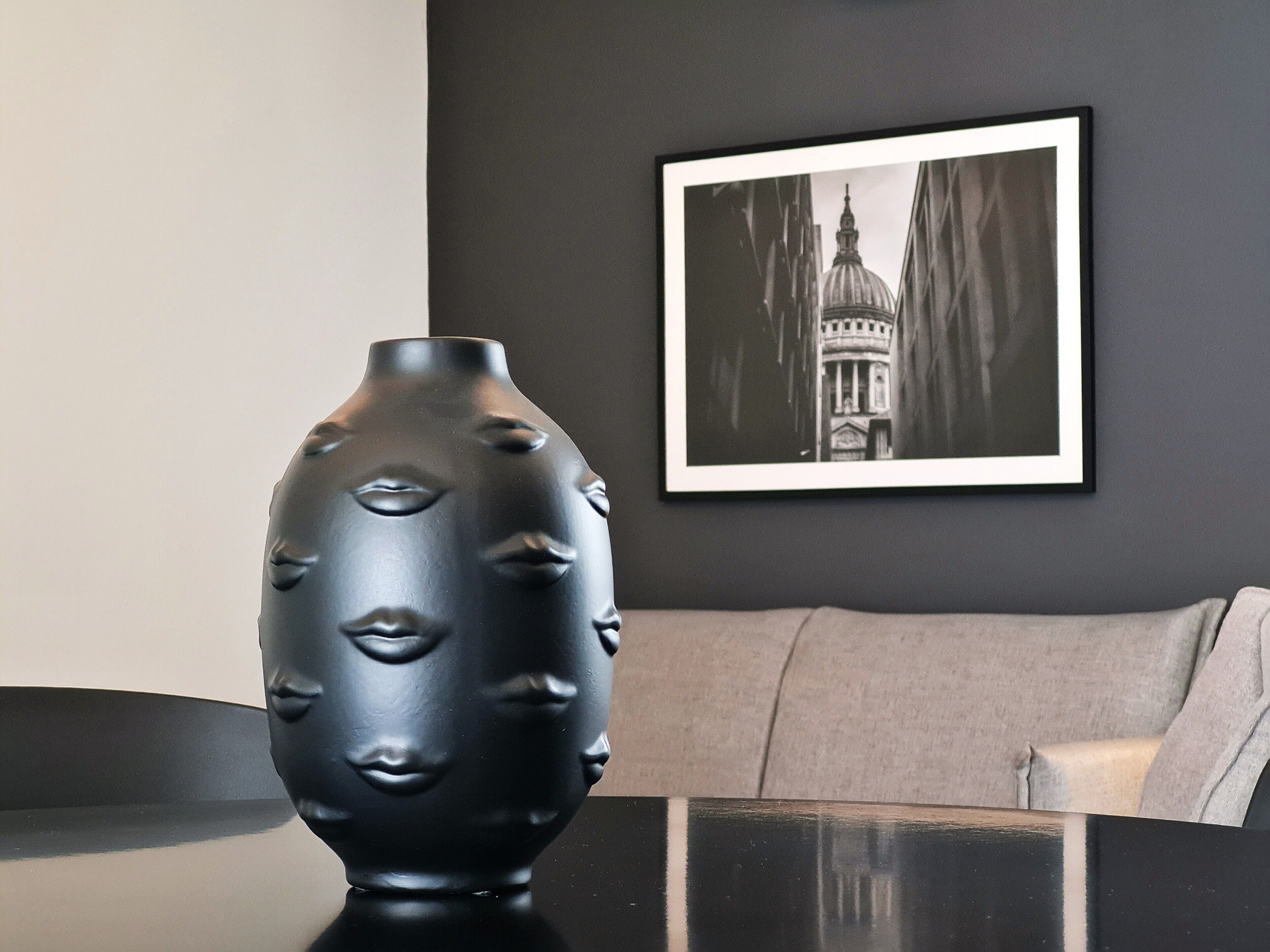 Eine schwarze Vase mit Gesichtszügen auf einem Tisch, ein gerahmtes Bild eines Gebäudes an der Wand und eine graue Couch im Hintergrund.