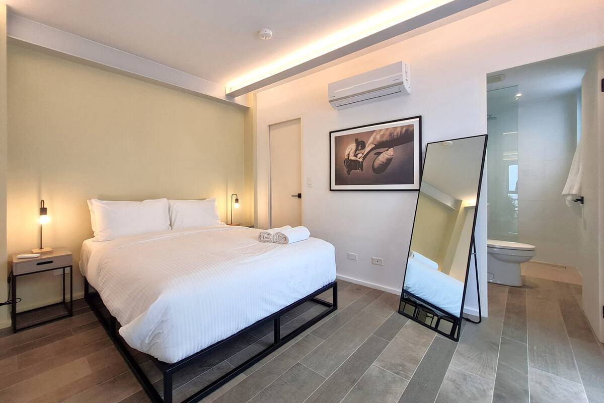 Ein modernes Hotelzimmer mit einem großen Bett, Nachttischlampen, einer Wand-Klimaanlage, einem gerahmten Kunstwerk und einem Badezimmer mit Toilette und Dusche.