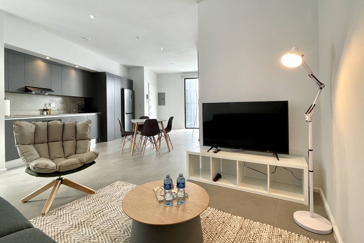 Ein modernes Wohnzimmer mit einer Couch, einem Couchtisch mit Wasserflaschen, einem Fernseher auf einem Schrank und einem Essbereich mit Stühlen und Tisch.