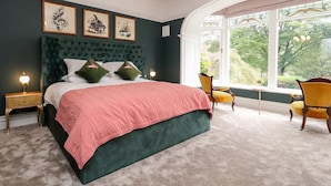 A bedroom with a large bed, two nightstands, a window with a view of greenery, and a chandelier.