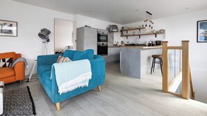 A modern living room with a blue sofa, an orange armchair, and a kitchen area with a bar counter. - La Casita - 2-bed House in Newquay (Newquay)