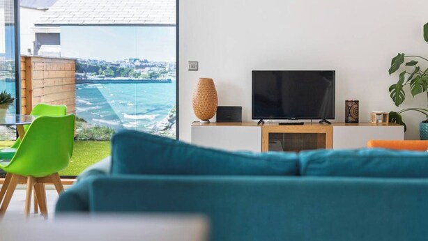 A modern living room with a large TV on a wooden stand, a green chair, and a view of the ocean through large windows. - La Casita - 2-bed House in Newquay (Newquay)