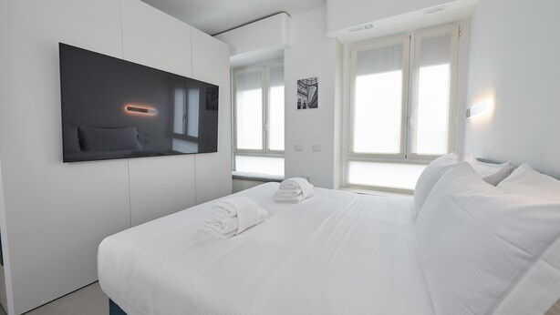 A modern hotel room with a large bed, a flat-screen TV mounted on the wall, and a large window providing natural light. - New apartment Central Station Milan (Milano)