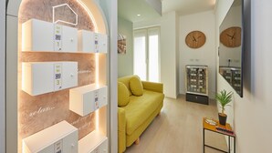 A modern living room with a yellow sofa, a small table with a plant, and a wall-mounted TV. There are two wooden clocks on the wall and a large arched window with illuminated storage units. - Circular Apartment . (Milano)
