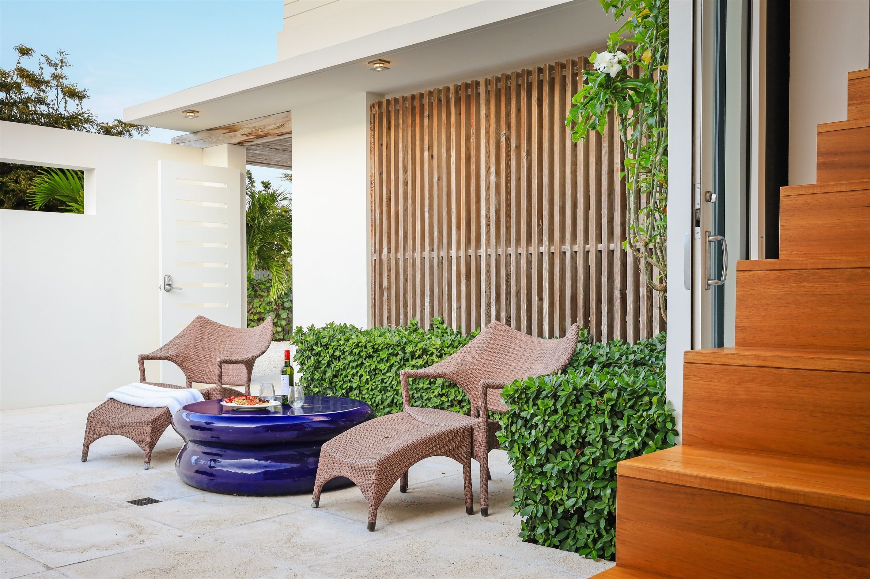 Un espace extérieur moderne, aménagé avec des chaises en osier et une table bleue, entouré de verdure et délimité par une clôture en bois.