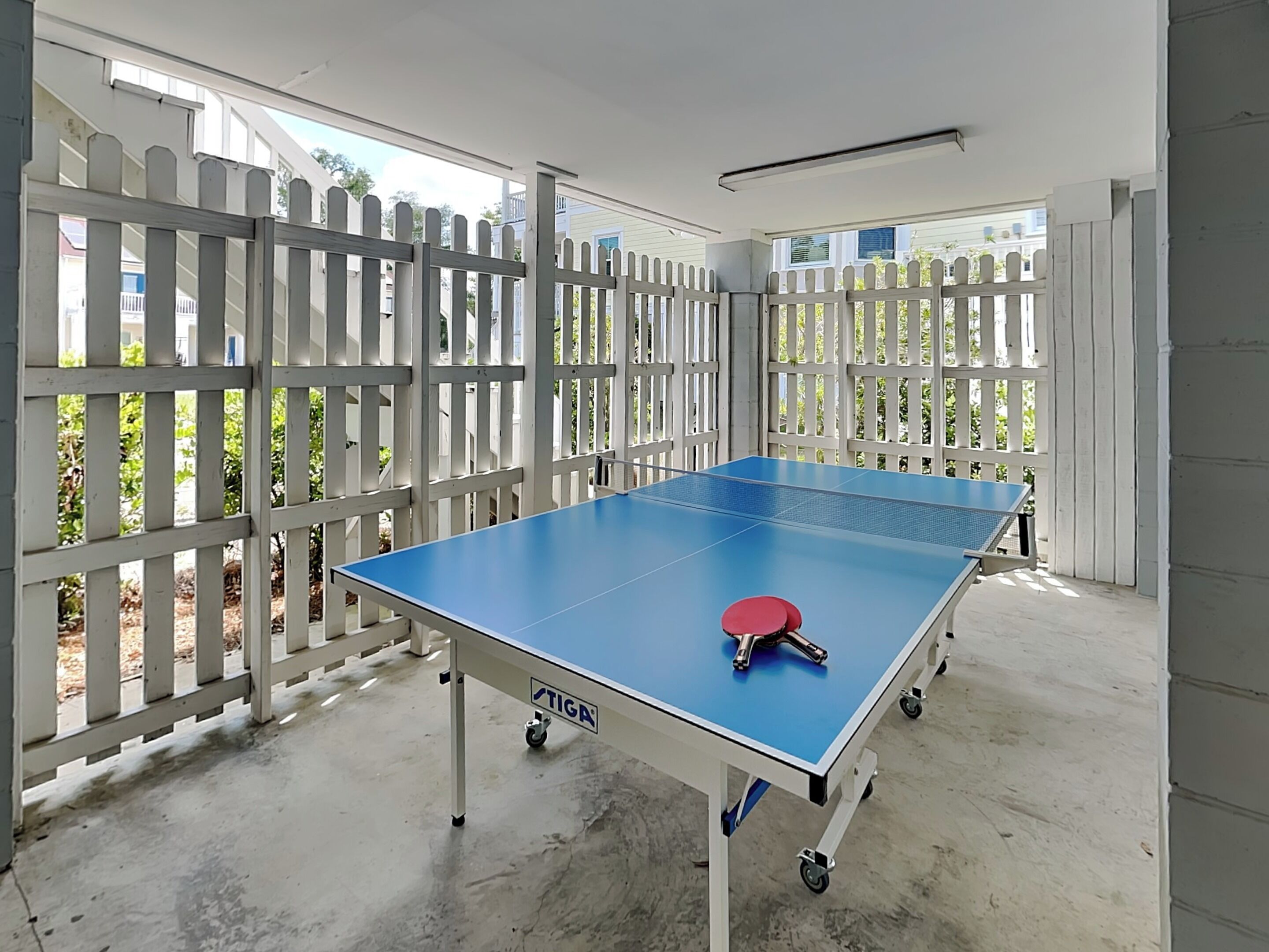 A ping pong table with wheels, a red paddle, and a 'STIGA' label, set against a white fence and a concrete floor.