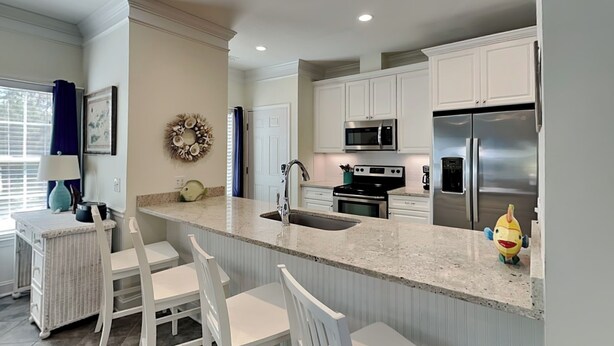 A modern kitchen with white cabinetry, stainless steel appliances, a granite countertop, and a breakfast bar with white chairs. - Spacious Tybee Retreat w/ Porch, Serene Setting Near Beach (Tybee Island)