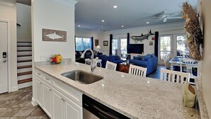 A modern kitchen with a dishwasher, a sink, and a dining area with blue chairs and a table. - Spacious Tybee Retreat w/ Porch, Serene Setting Near Beach (Tybee Island)