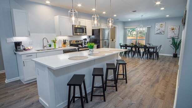 A modern kitchen with a central island, bar stools, and a dining area with a table and chairs. - Escape to 4br/3ba Brand New Downtown Home (San Antonio)