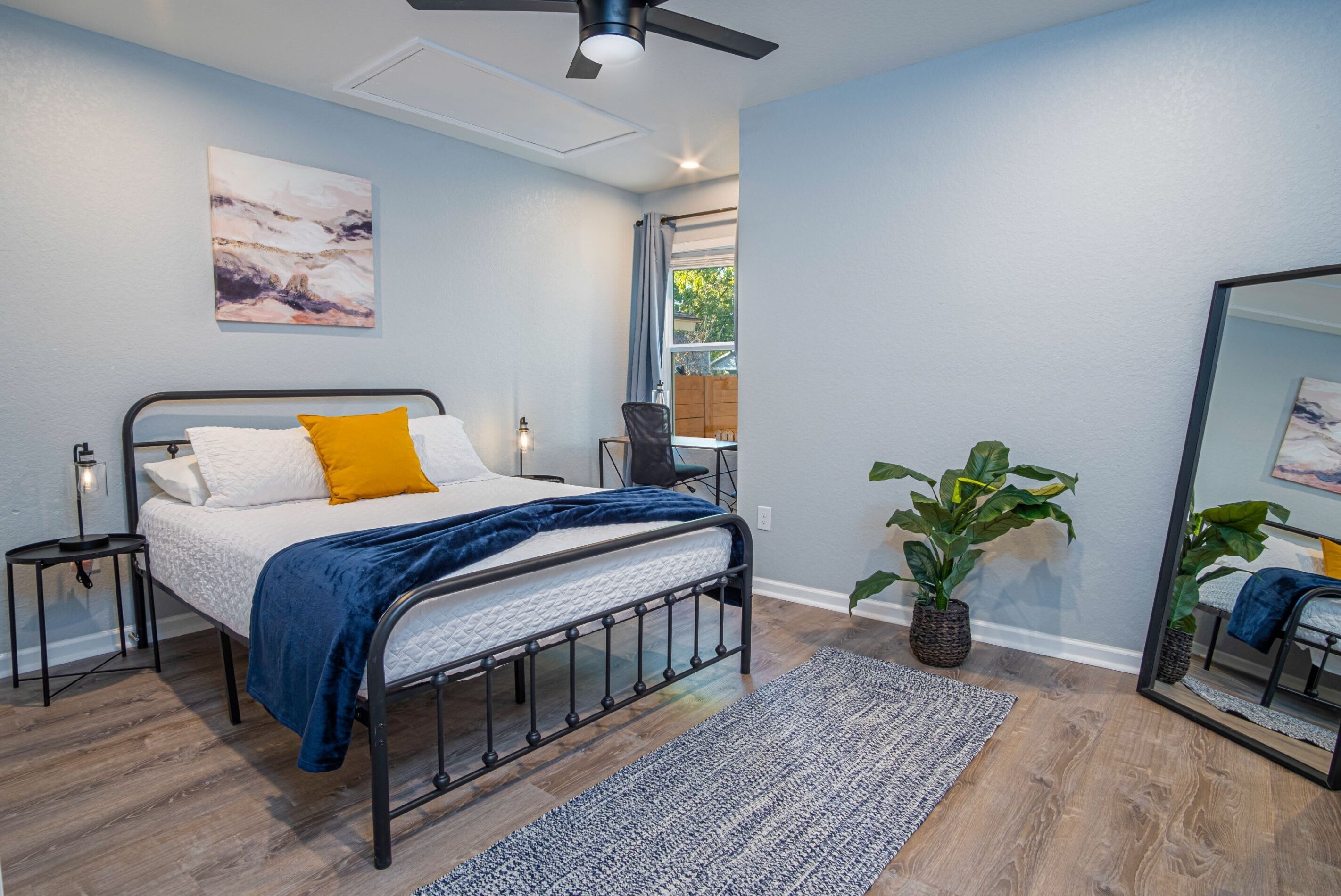 A bedroom with a bed, bedside tables, a ceiling fan, a large window, and a potted plant.