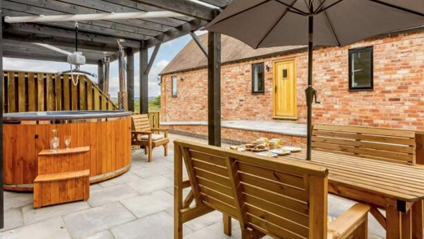 An outdoor patio area with wooden furniture, a barbecue grill, and a large umbrella. - Fawn Cottage Disabled Adapted 3 Bedroom hot tub (Alcester)