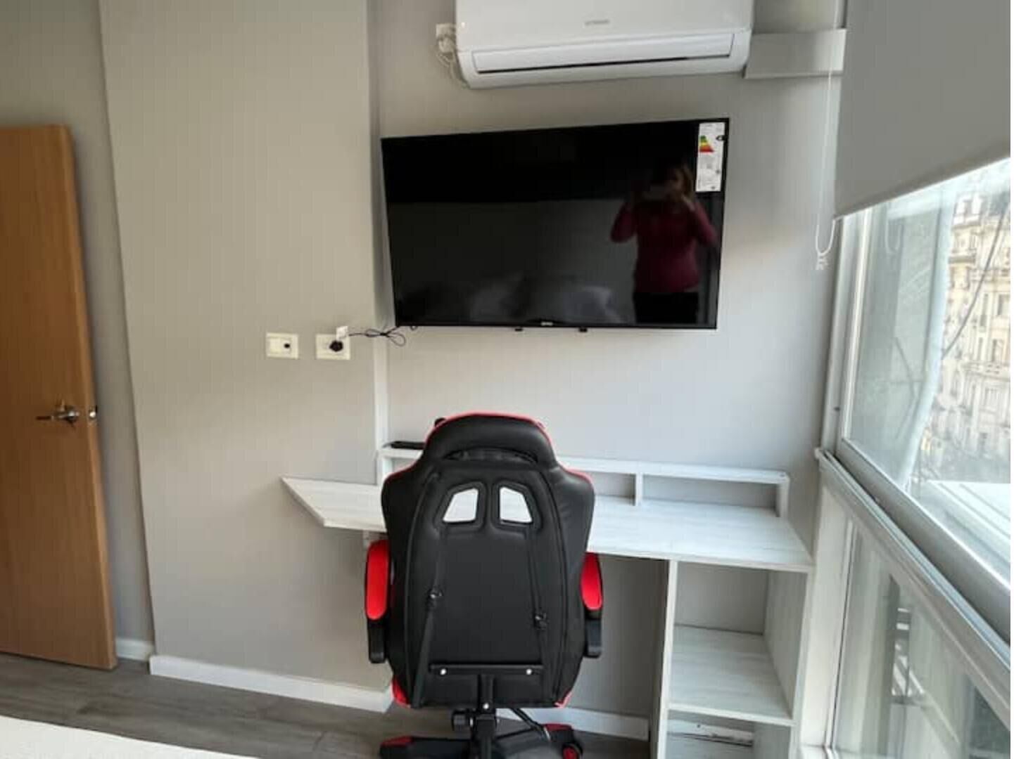 Ein Zimmer mit einem an der Wand montierten Fernseher, einem Gaming-Sessel und einem Fenster mit Blick auf Gebäude.