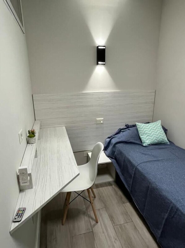 Une petite chambre à coucher moderne, avec un lit, un bureau et une chaise. Il y a une lampe fixée au mur au-dessus du lit et une petite plante sur le bureau.