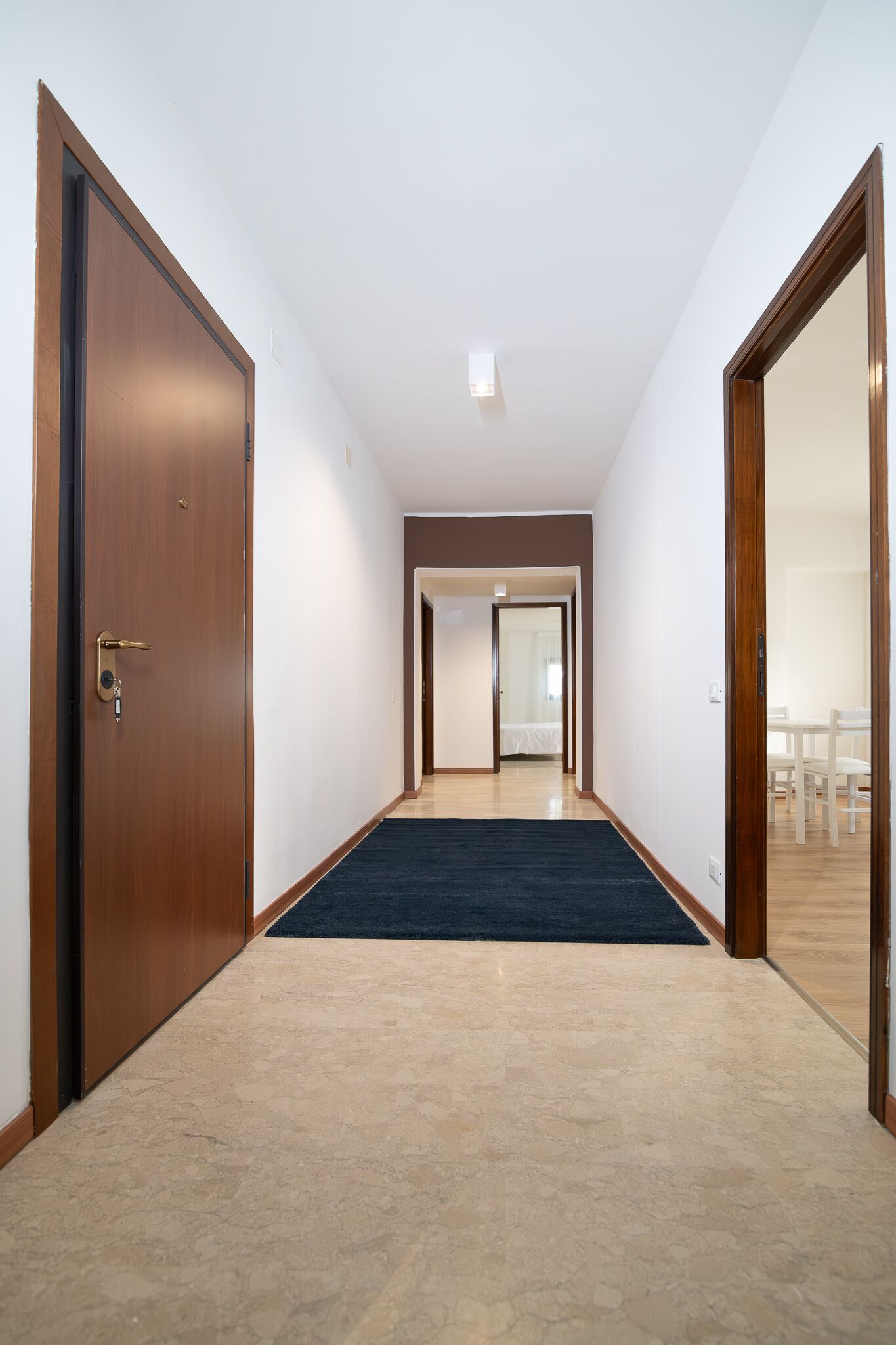 A hallway with a carpeted floor, wooden doors, and a view into a room with a bed and a table.