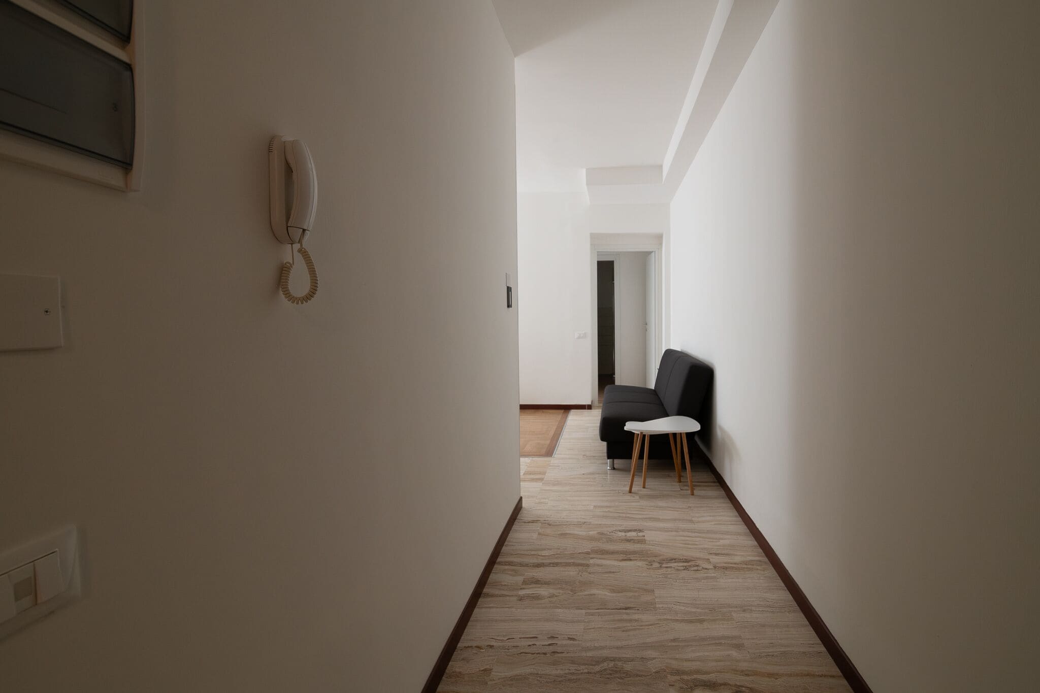 A narrow hallway with a white wall, a telephone mounted on the left, and a dark-colored sofa with a small white table in the distance.
