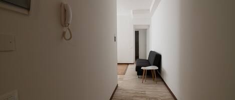A narrow hallway with a white wall, a telephone mounted on the left, and a dark-colored sofa with a small white table in the distance.