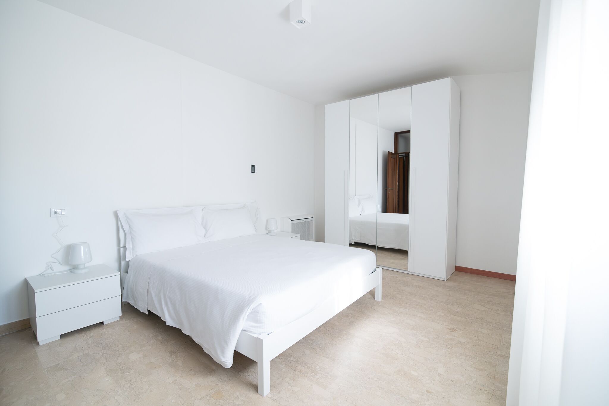 A minimalist bedroom with a large bed, a nightstand, a wardrobe, and a mirror.