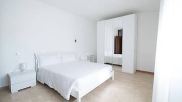 A minimalist bedroom with a large bed, a nightstand, a wardrobe, and a mirror.
