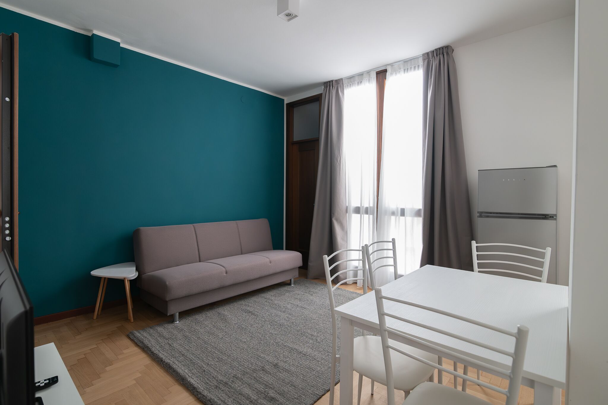 Un salon moderne avec des murs de couleur turquoise, un canapé gris, une petite table blanche et un coin repas avec des chaises blanches.