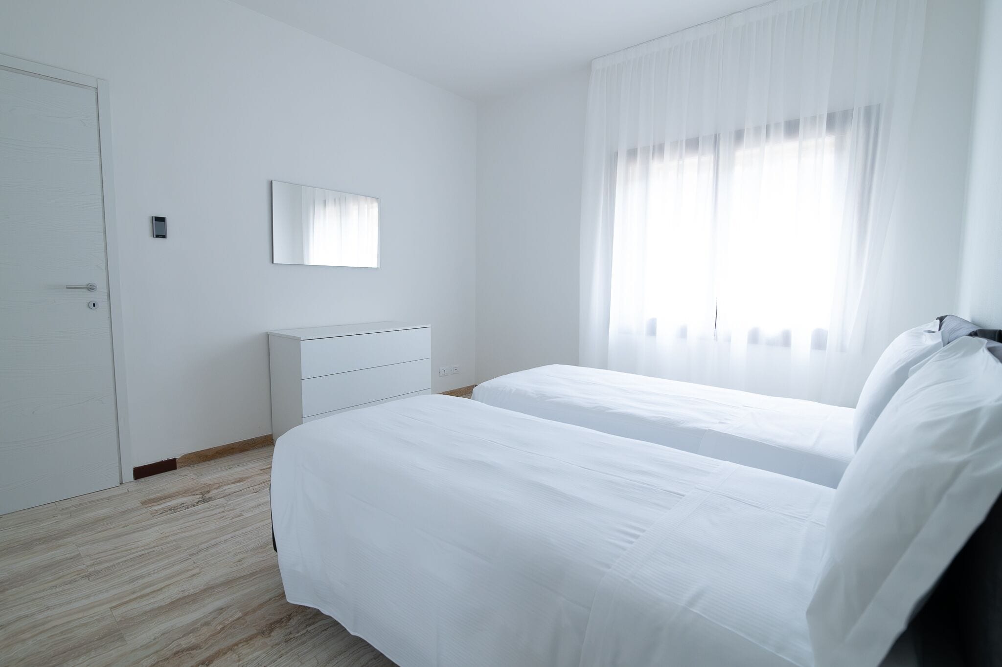 Une chambre à coucher au style minimaliste, avec deux lits, une commode blanche, un miroir et une porte.