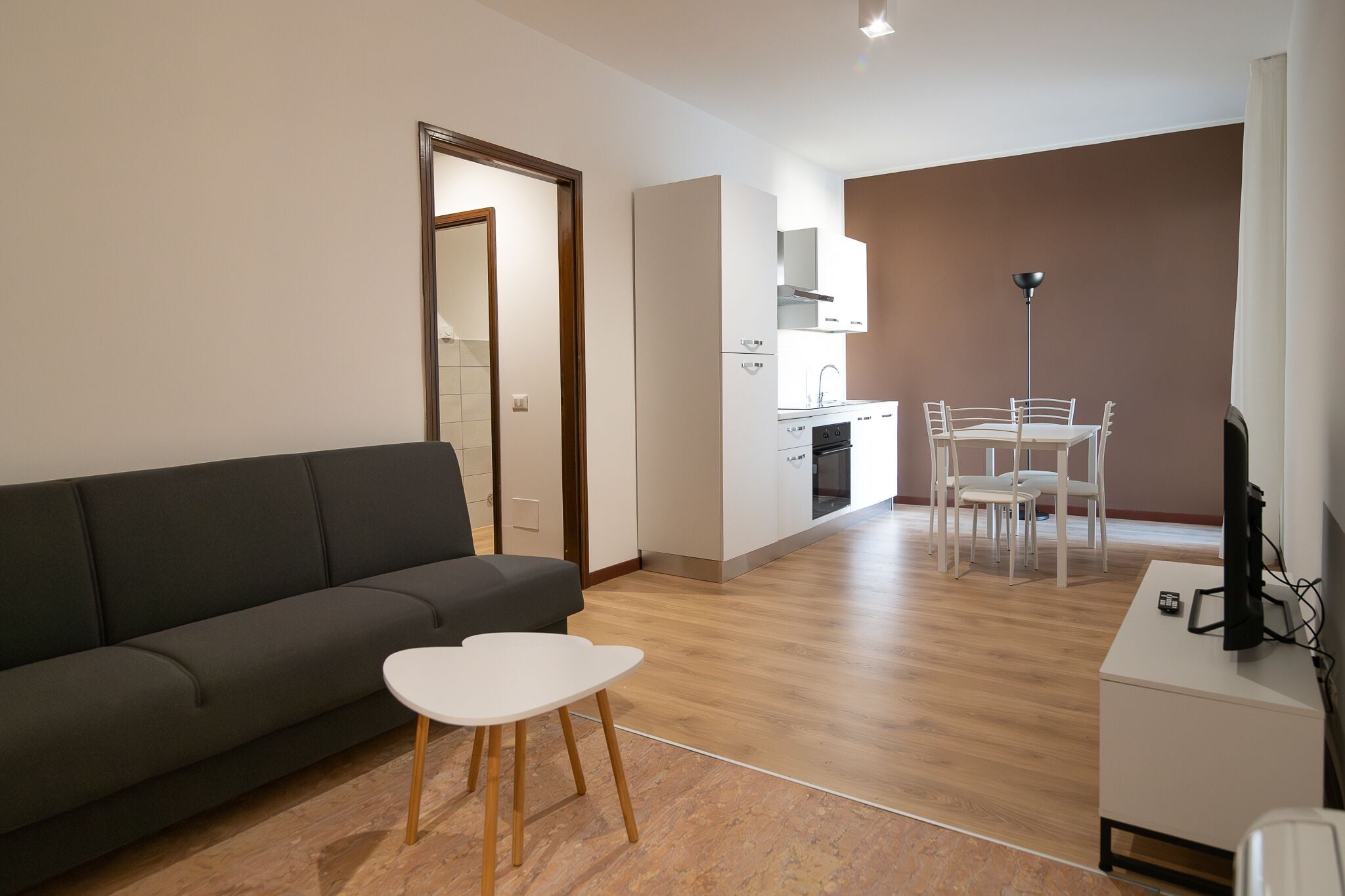 A modern living room with a sofa and a small table, a dining area with chairs, a kitchen with cabinets, and a TV on a stand.