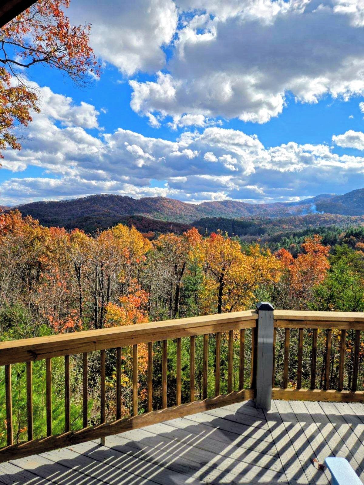 A wooden deck with a railing overlooking a forested valley with autumn foliage and distant mountains.