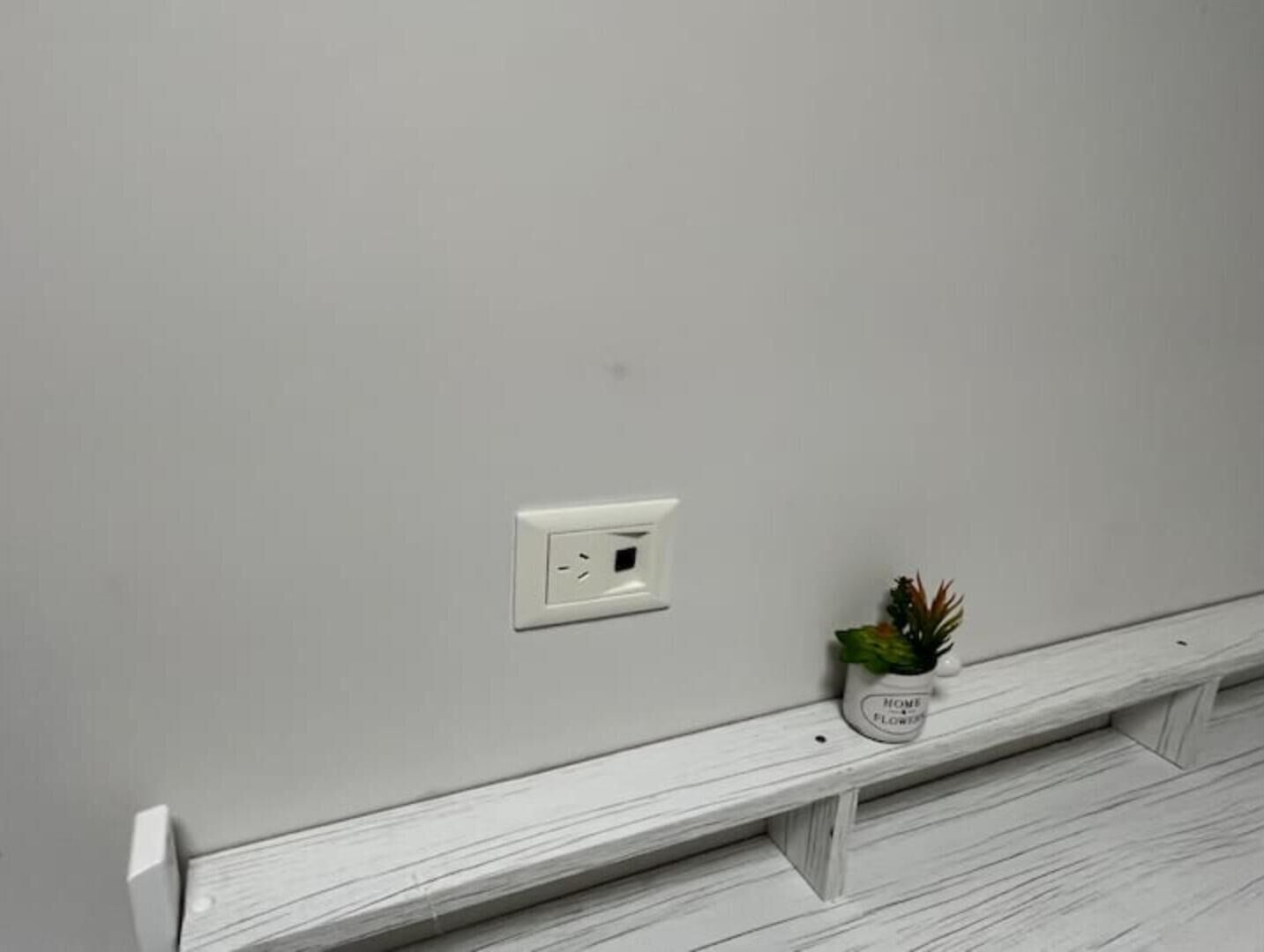 A white wall with a power outlet and a small potted plant on a white shelf.