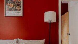 A room with a red wall, a white sofa with patterned cushions, a floor lamp, and a framed picture on the wall. - Kensington Stay (London)