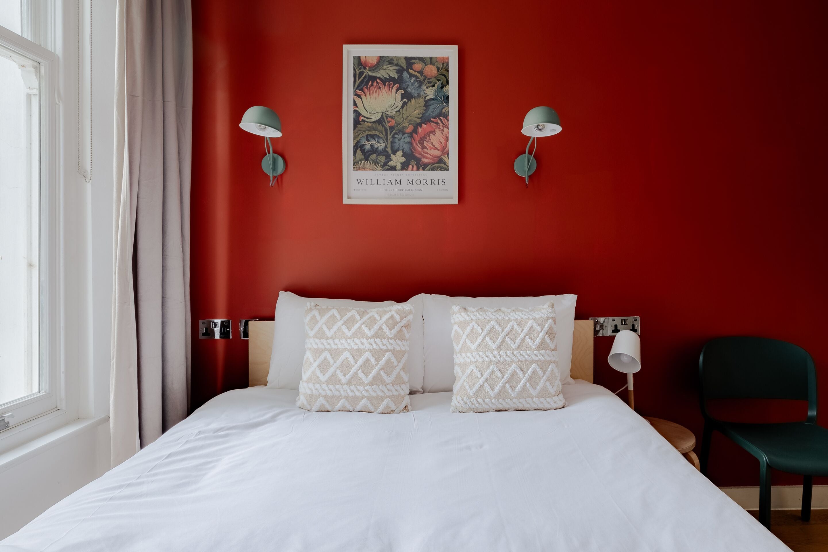 A bedroom with a red wall, white bedding, two patterned pillows, a framed picture, and two wall-mounted lamps.
