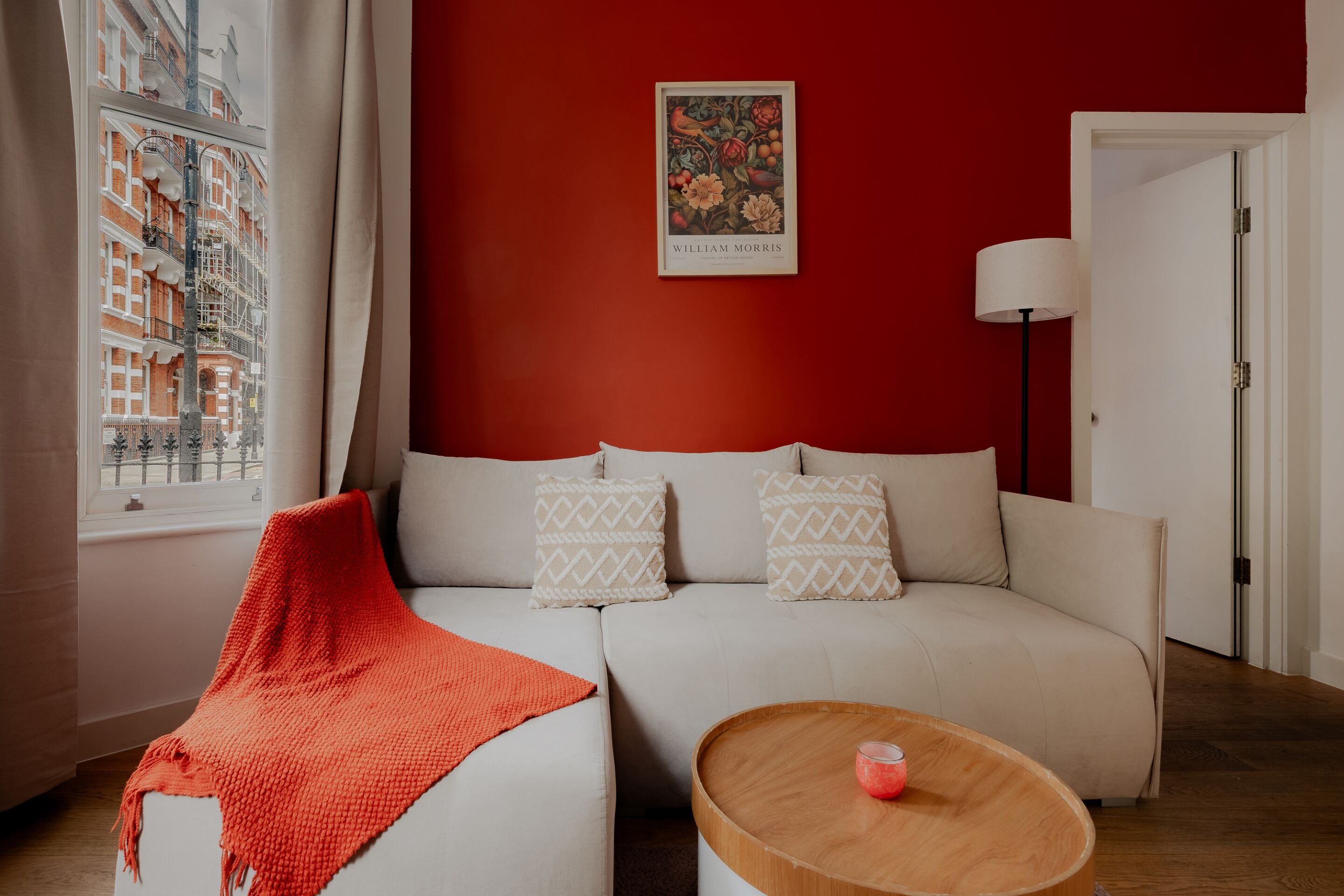 A modern living room with a red accent wall, a white sofa with patterned cushions, a red throw blanket, and a wooden coffee table with a glass candle.