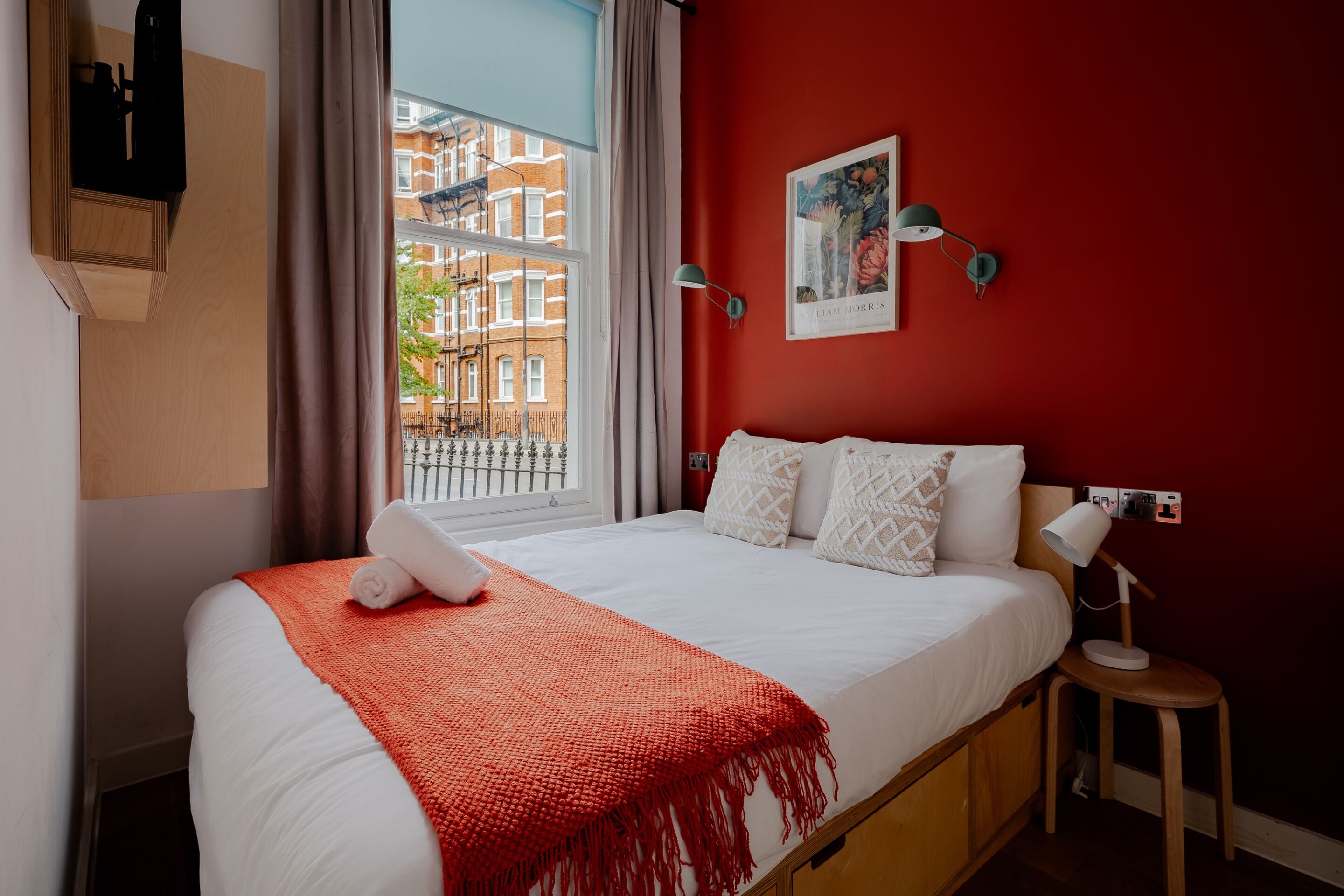 A hotel room with a large bed, a red wall, a window with a view of buildings, and a framed picture on the wall.