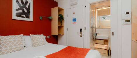 Une chambre d’hôtel avec un lit, un mur rouge, une œuvre d’art abstraite encadrée, une salle de bain avec un lavabo et un miroir, et une porte avec une pancarte.
